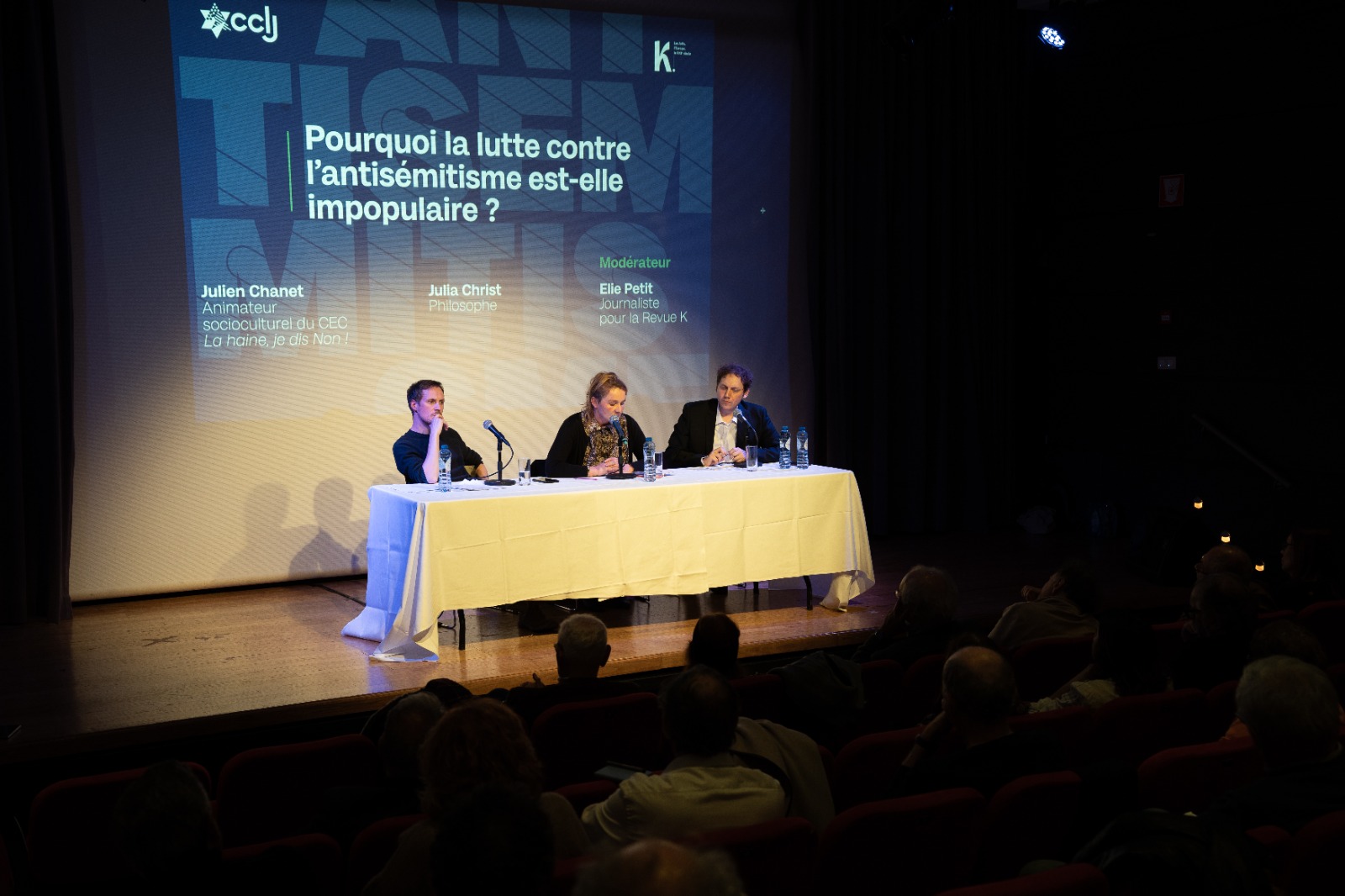 Trois intervenants assis derrière une table blanche sur une scène face à un public dans une salle de conférence. Derrière eux, un grand écran projette le titre de la rencontre : « Pourquoi la lutte contre l’antisémitisme est-elle impopulaire ? », ainsi que les noms des participants et du modérateur.