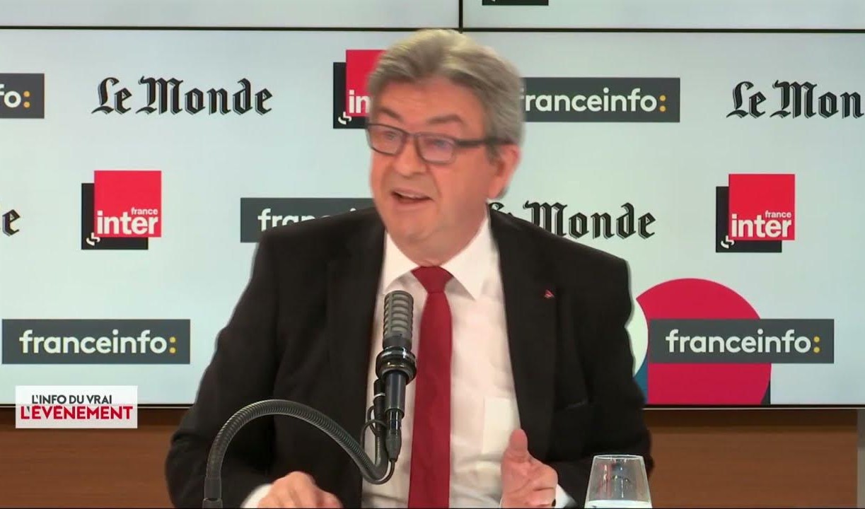 Mélenchon en costume noir et cravate rouge parle devant un microphone dans un studio d’interview ou de débat. Derrière lui apparaissent sur un écran les logos de France Inter, franceinfo et Le Monde.