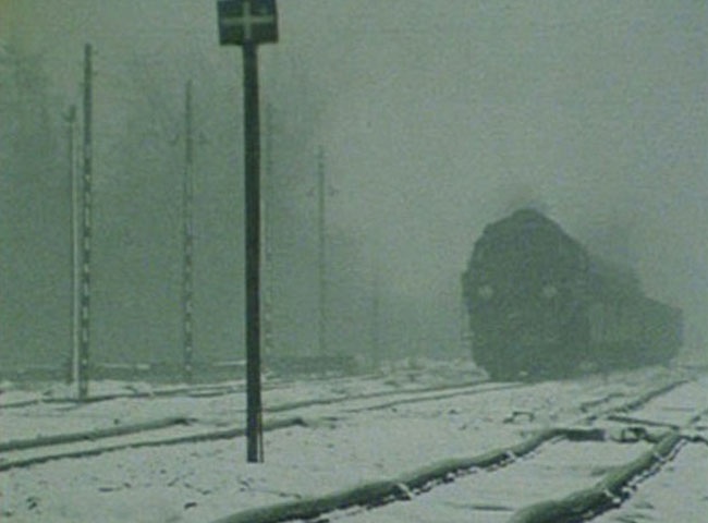 Image granuleuse montrant une locomotive sombre avançant sur des voies ferrées enneigées dans un paysage brumeux. Plusieurs poteaux bordent les rails, tandis que le train apparaît partiellement enveloppé de fumée ou de brouillard.