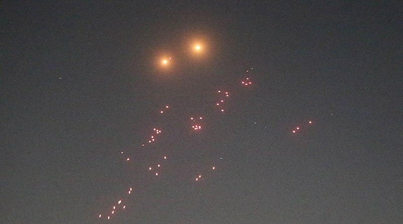 Ciel nocturne sombre traversé par de multiples points lumineux et traînées incandescentes dispersés dans différentes directions. Deux sources lumineuses plus intenses brillent dans la partie supérieure de l’image, tandis que de nombreux éclats rouges et orangés parsèment le ciel.