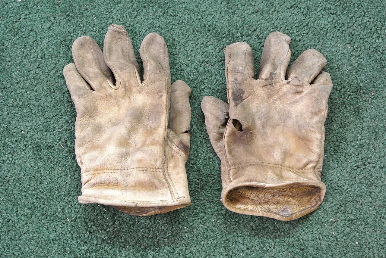 Deux gants en cuir beige fortement usés et tachés, posés à plat sur une surface textile verte. Le gant de droite présente plusieurs déchirures et trous visibles au niveau du dos de la main et près du poignet.