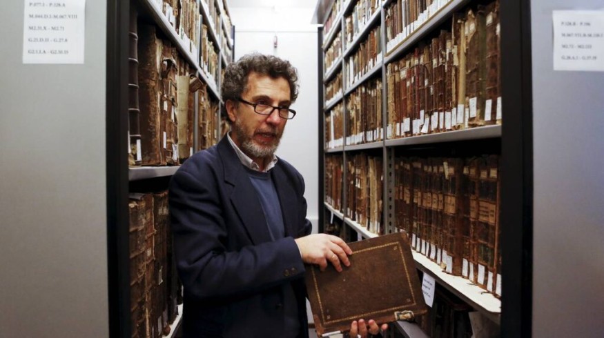 Photographie de Gadi Luzzatto Voghera debout dans une allée étroite entre deux rayonnages d’archives remplis de livres anciens reliés en cuir. Il tient un volume ancien dans les mains et regarde légèrement hors champ. Il porte des lunettes et une veste sombre.