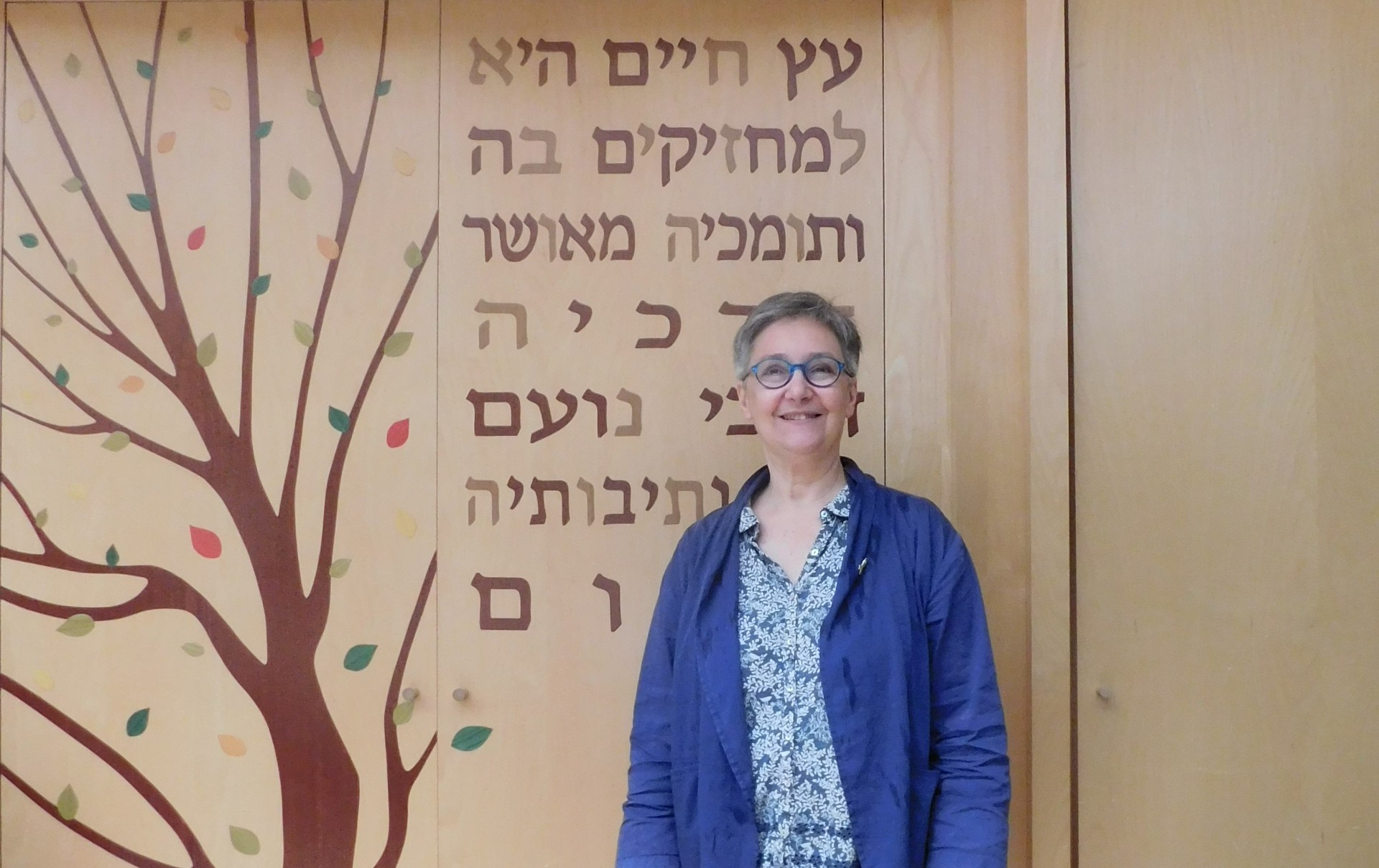 Portrait d’une femme souriante portant des lunettes et une veste bleue, debout devant un mur en bois clair décoré d’un arbre stylisé et de plusieurs lignes de texte en hébreu.
