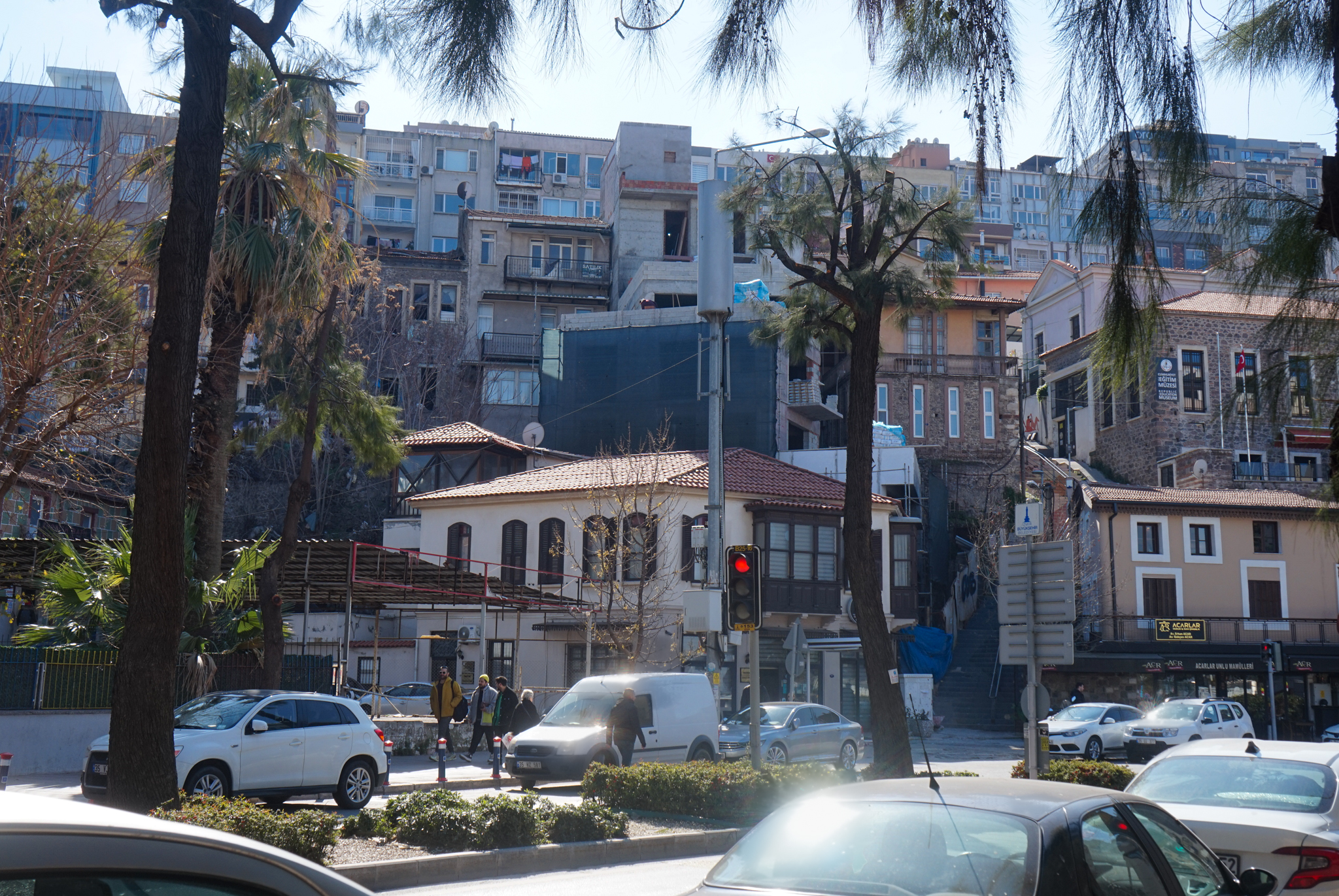 Vue urbaine du quartier de Karataş à Izmir, ancien quartier juif de la ville, montrant des maisons basses traditionnelles au pied d’immeubles modernes sur une colline, bordées par une avenue avec circulation et arbres au premier plan.
