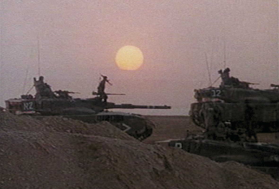 Image au grain cinématographique montrant plusieurs chars israéliens sur un terrain désertique au coucher ou lever du soleil. Un soldat israélien se tient debout sur l’un des chars tandis qu’un large soleil apparaît bas dans le ciel à l’arrière-plan.