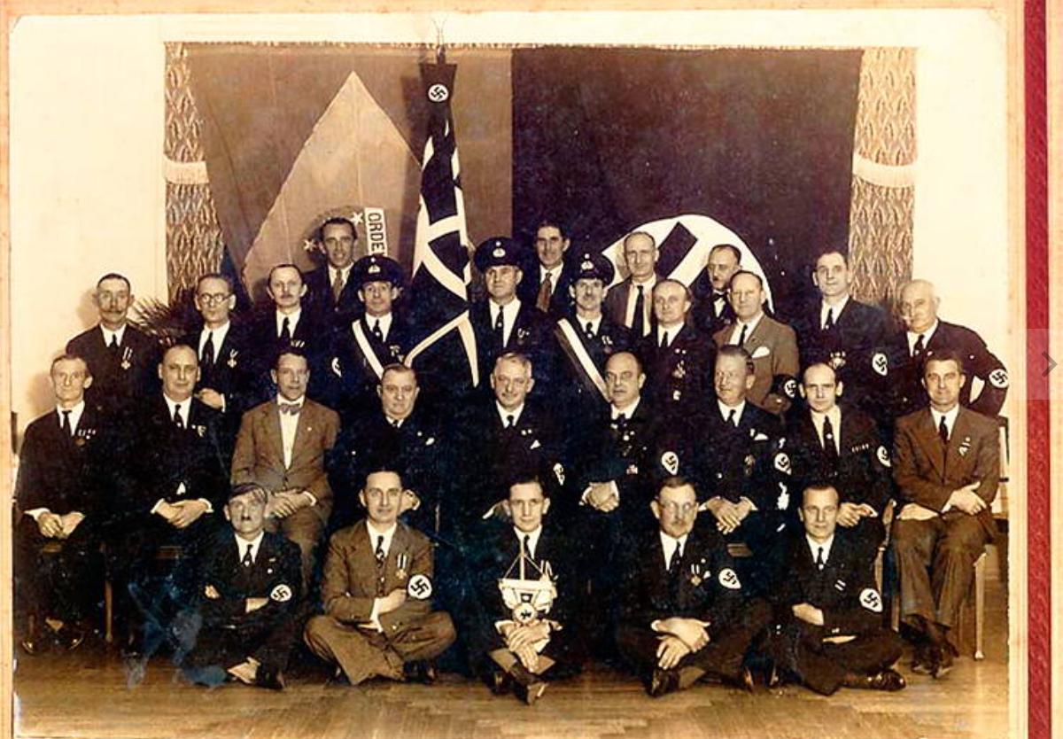 Photographie de groupe en intérieur montrant plusieurs dizaines d’hommes posant en rangs dans une salle décorée de drapeaux nazis et brésiliens. Certains sont assis au premier rang, d’autres debout derrière eux, plusieurs portent des brassards à croix gammée ou des uniformes paramilitaires. Au centre, derrière le groupe, de grands emblèmes nazis et une bannière verticale ornée d’une croix gammée dominent la scène. L’image est composée comme un portrait officiel de rassemblement politique, avec tous les participants tournés vers l’objectif.