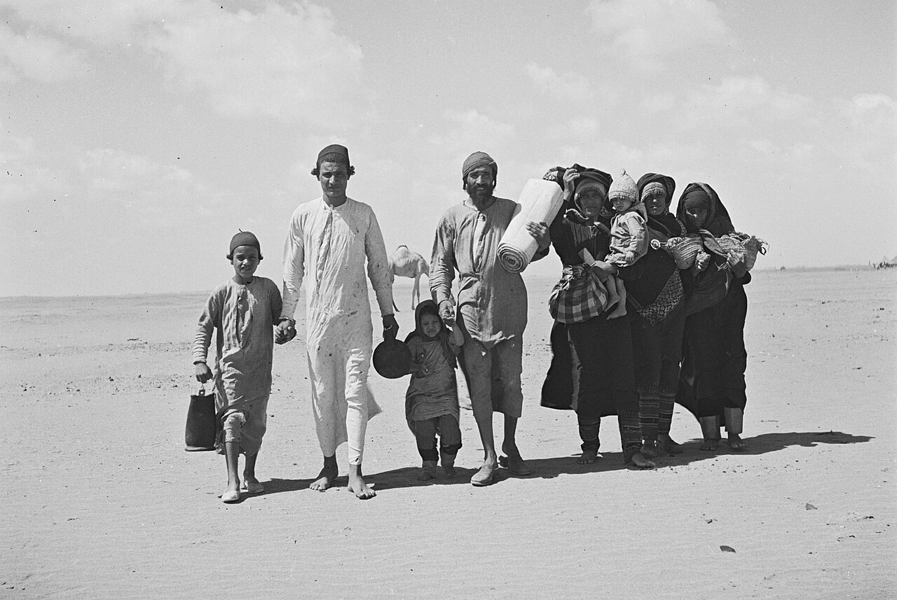 Photographie d’archive en noir et blanc montrant une famille de Juifs yéménites marchant à travers une étendue désertique en 1949 en direction d’un camp sioniste à Aden avant leur départ vers Israël, composée d’hommes, femmes et enfants portant ballots, couvertures et nourrissons, dans une scène de migration collective capturant les conditions précaires de l’exode des communautés juives du Yémen au lendemain de la création de l’État d’Israël.