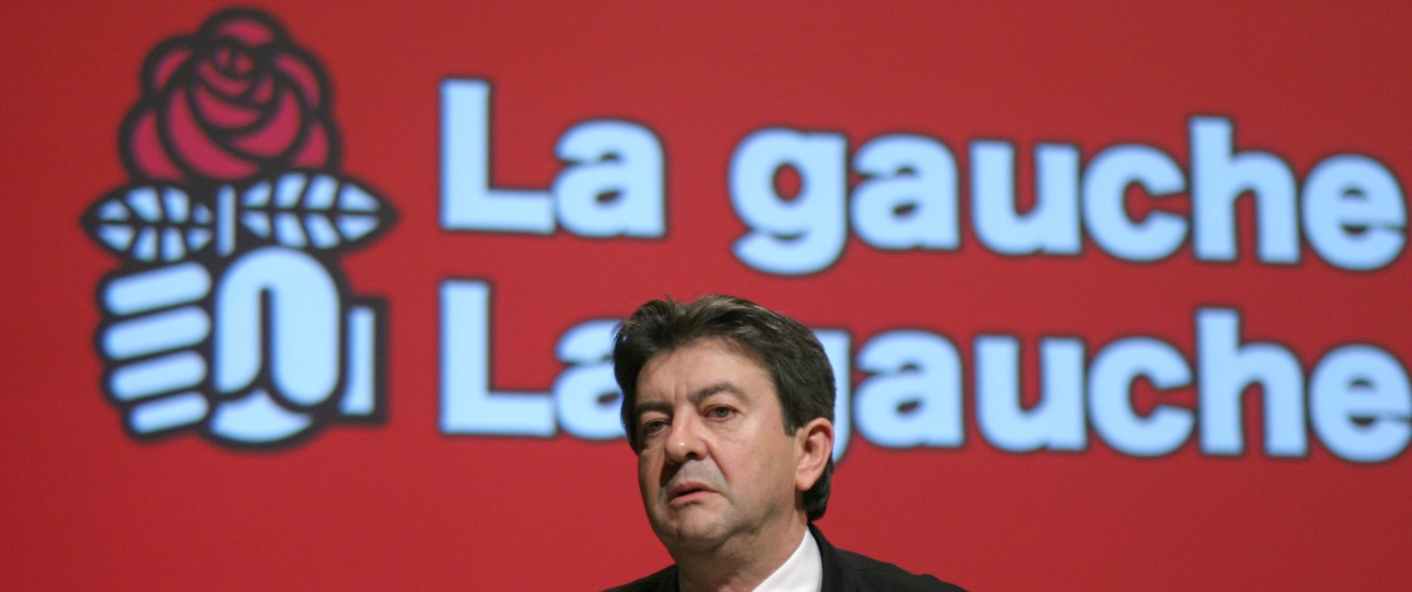 Photographie de Jean-Luc Mélenchon s’exprimant derrière un pupitre lors d’un meeting ou congrès politique du Parti Socialiste, vêtu d’un costume sombre et d’une cravate, devant un large écran rouge affichant le logo à la rose et « La gauche La gauche » écrit en blanc en arrière-plan.