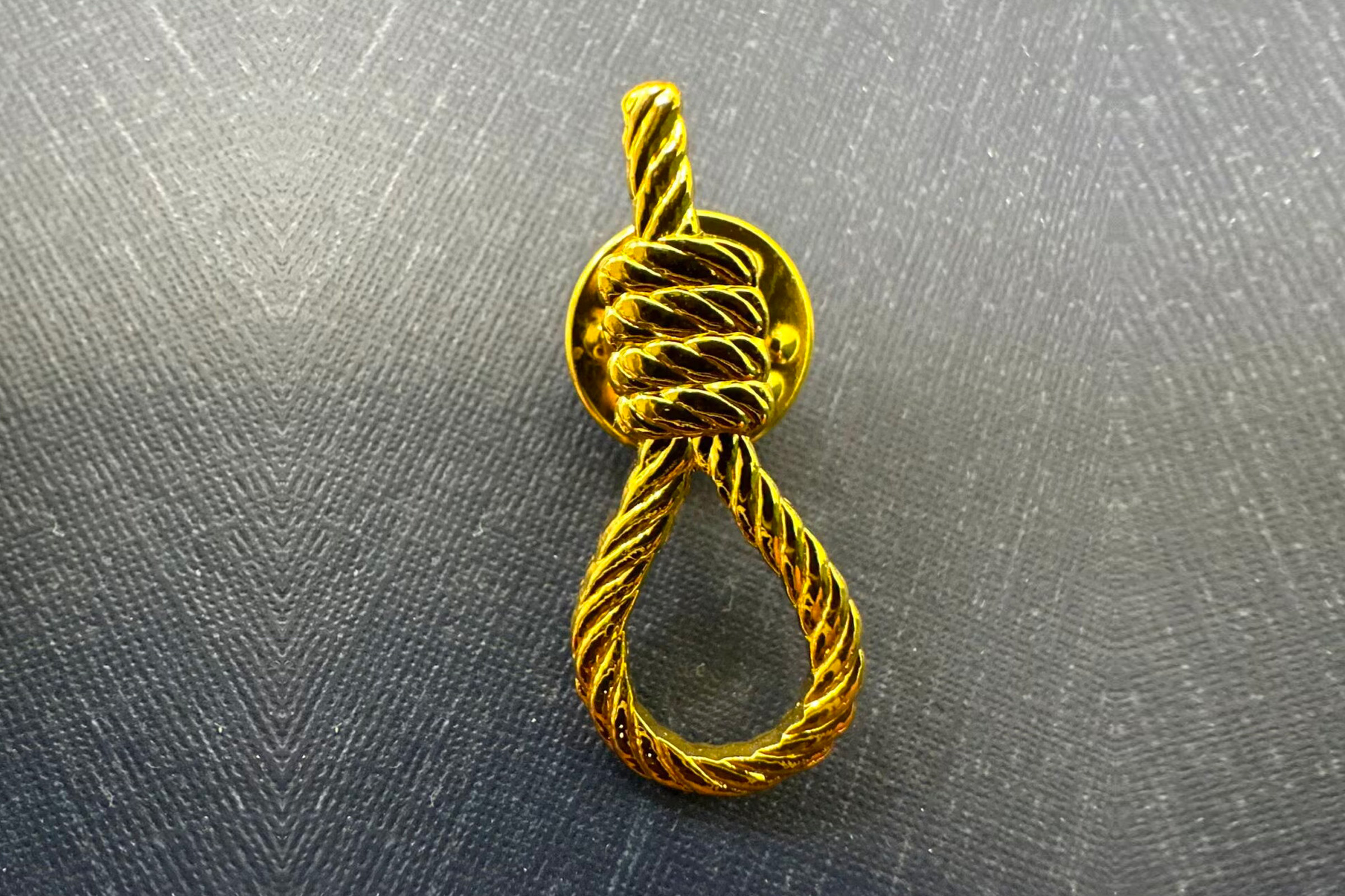 Photographie en gros plan d’une broche ou épinglette dorée représentant un nœud coulant stylisé en corde torsadée, posée sur un fond texturé gris.