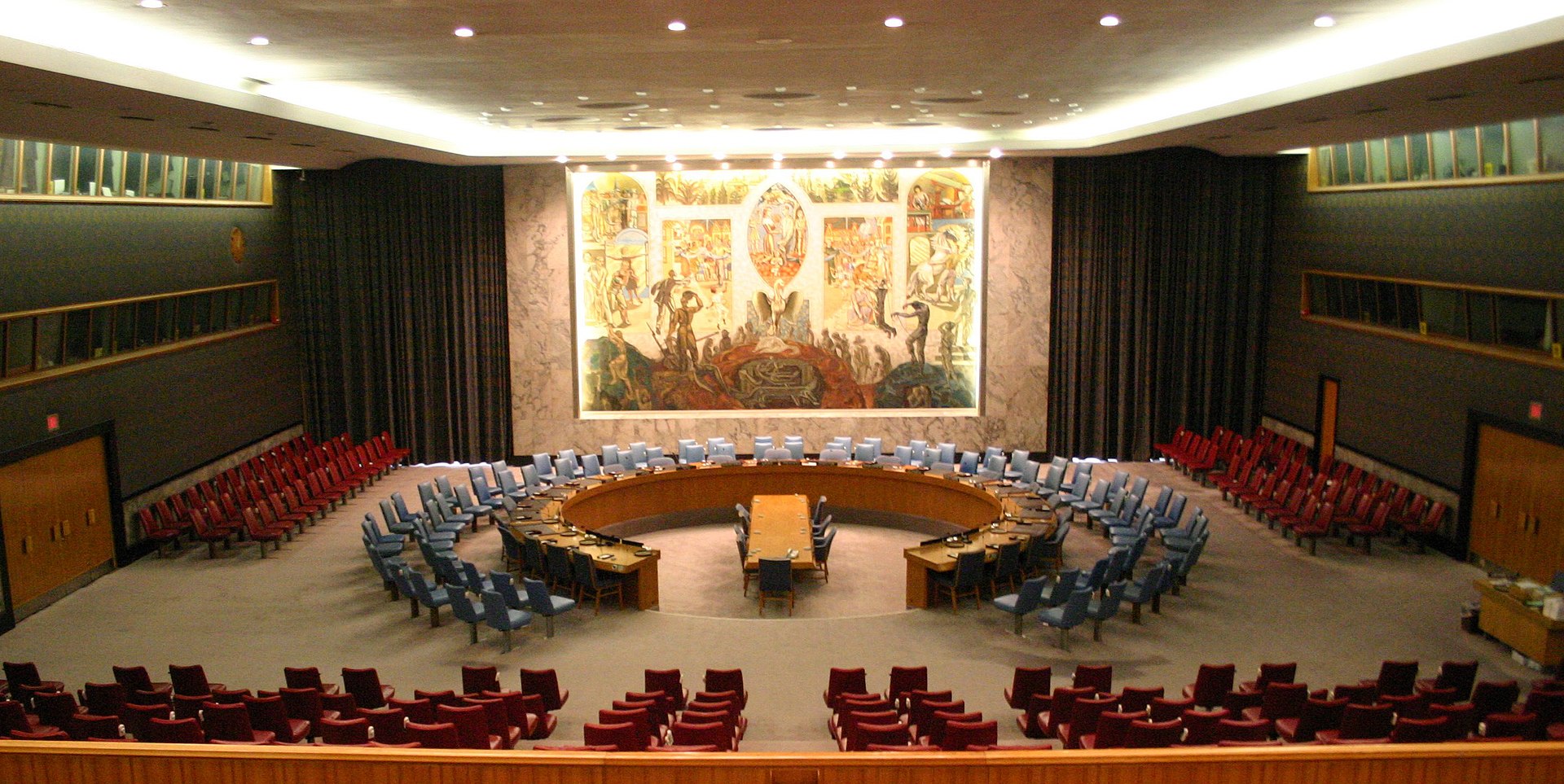 Photographie d'une panoramique de la salle du Conseil de sécurité des Nations unies au siège de l’ONU à New York, montrant la table circulaire des délégations diplomatiques, les rangées de sièges pour observateurs et la grande fresque murale emblématique en arrière-plan.