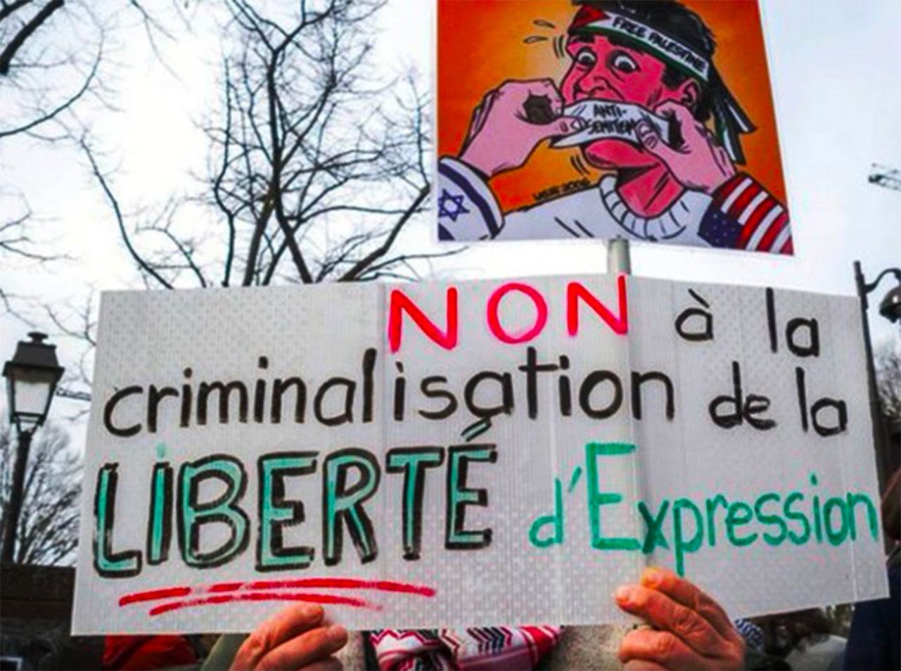 Manifestant tenant une grande pancarte portant le message « Non à la criminalisation de la liberté d’expression ». Au-dessus, une autre affiche illustrée représente un personnage caricatural avec un bandeau sur fond orange, dans un contexte de rassemblement en extérieur.