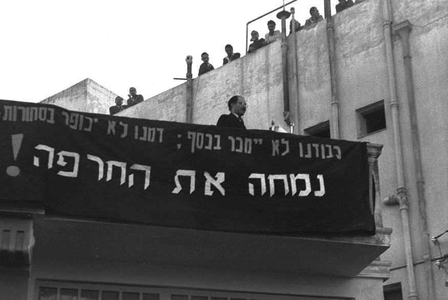 Manifestation de 1952 en Israël contre tout accord avec l'Allemagne avec sur scène Menahem Begin. Le panneau indique : « Notre honneur ne sera pas vendu pour de l'argent ; notre sang ne sera pas expié par des biens. Nous effacerons la honte ! ». Licence Wikimedia Commons